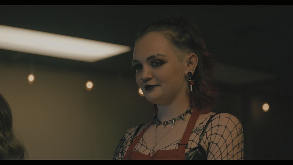 A young woman in a spiked choker and red apron smiles under fluorescent light