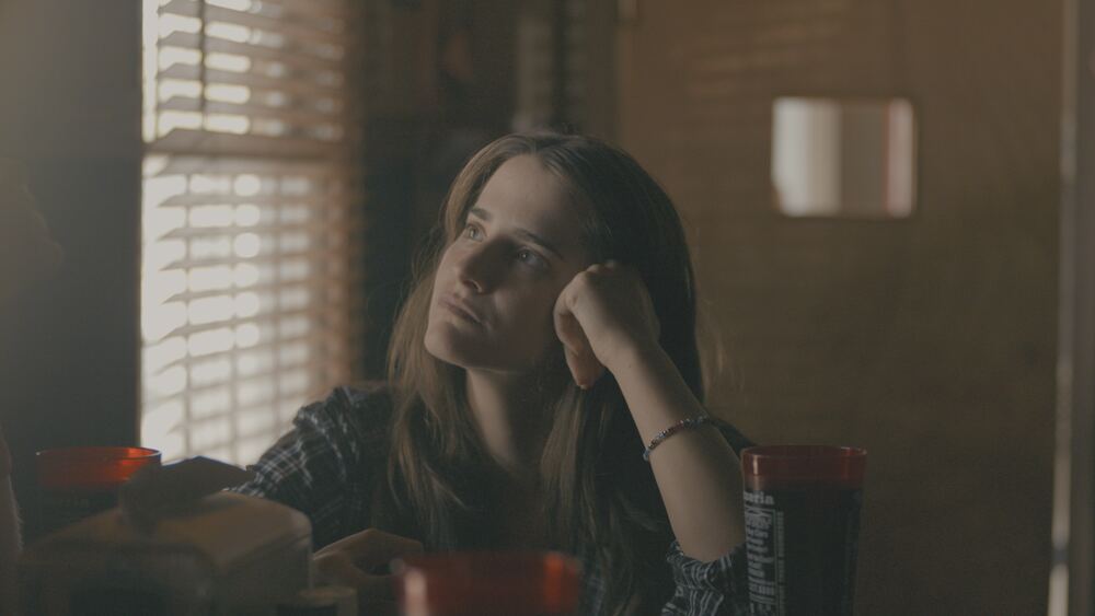 A young woman rests her head on her hand at a diner table by window blinds